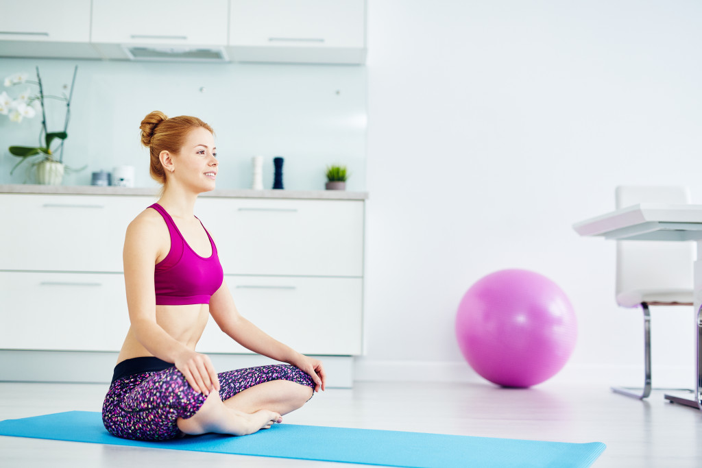 Portrait of fit red haired woman doing yoga exercises at home on floor: sitting with legs crossed in lotus position on mat and smiling